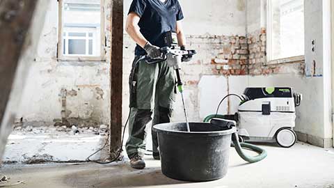 A craftswoman is standing on a construction site using a Festool mixer in combination with a dust extractor.